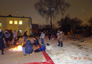Dzieci z rodziacami pieką kiełbaski przy ognisku.